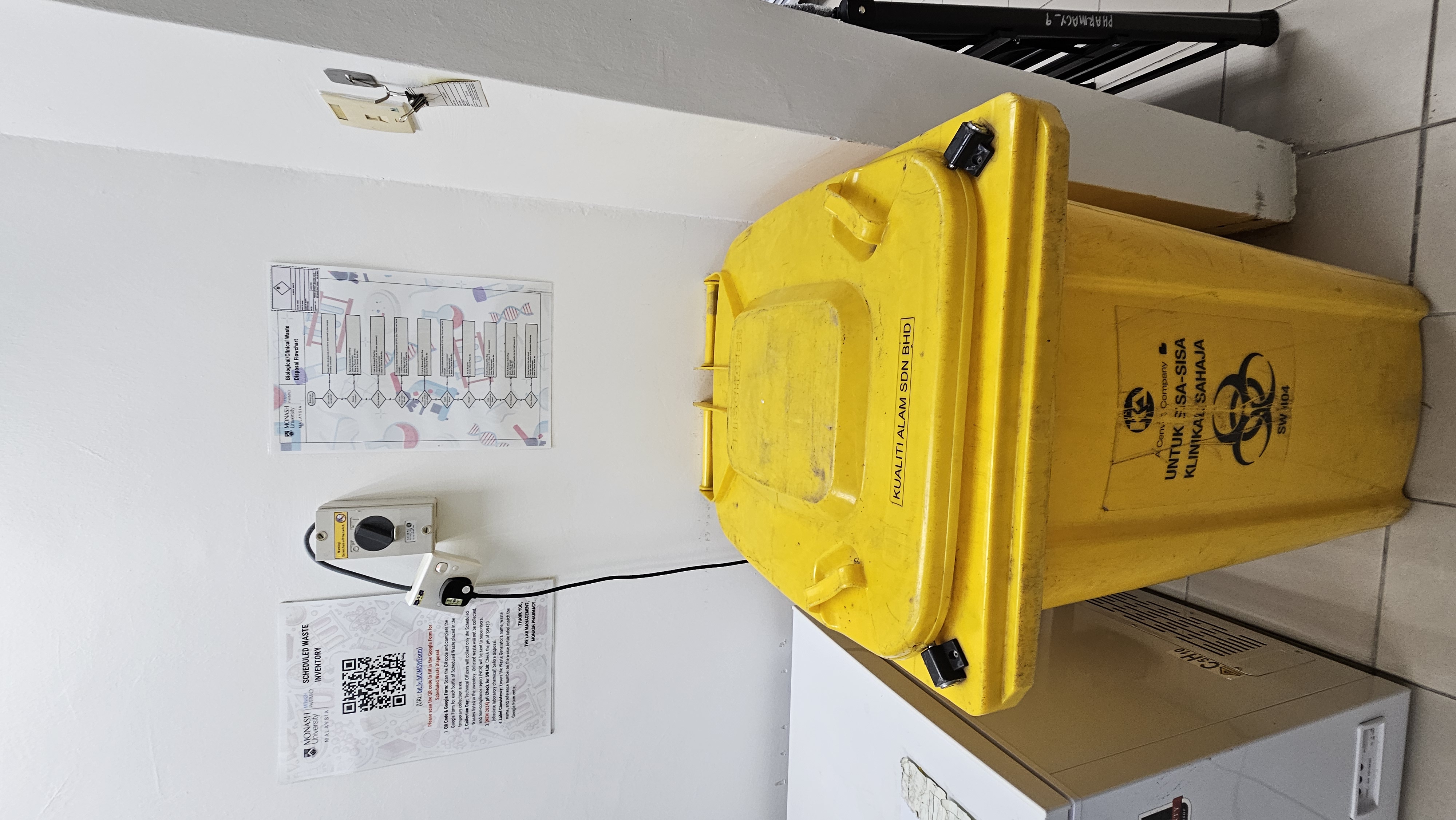 The wall above the yellow clinical waste bin at 3-6-17A: the Scheduled Waste Inventory QR poster on the left and the Biological/Clinical Waste Disposal Flowchart on the right