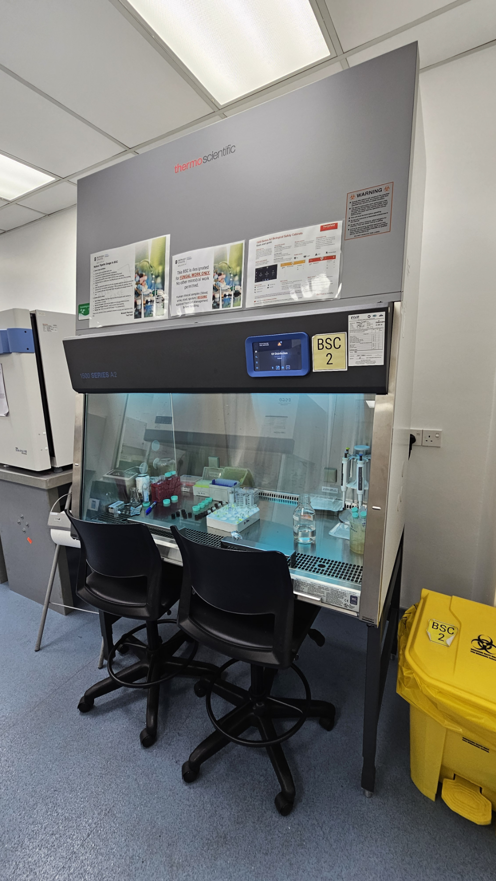 Thermo Scientific 1500 Series A2 biosafety cabinet running UV disinfection, with usage notices above the sash and a yellow BSC 2 clinical waste bin beside it