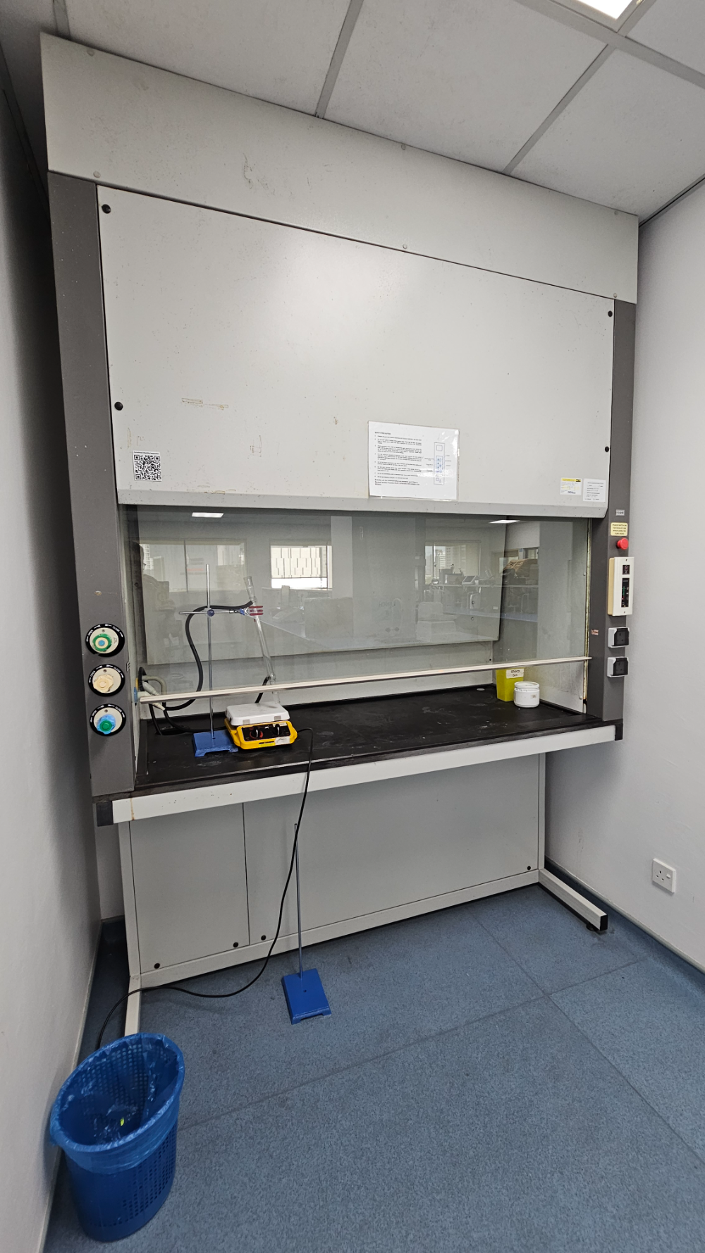 Walk-in fume hood in the lab with the sash lowered, safety precaution notice on the front, gas and water service taps at the side, and a small sharps bin on the deck
