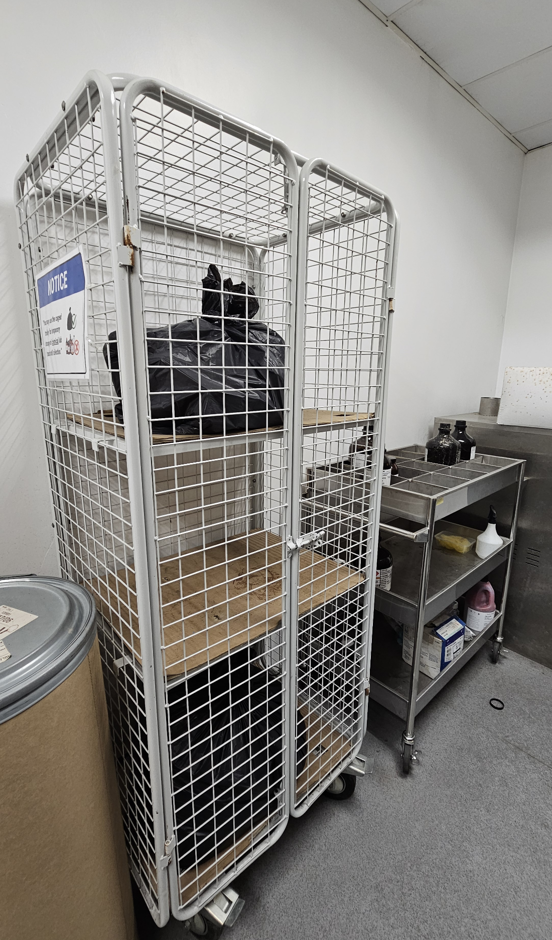 Caged trolley inside 4-6-09 holding tied black bags of chemical lab trash, with a notice on the cage, and a stainless steel trolley beside it holding empty chemical bottles