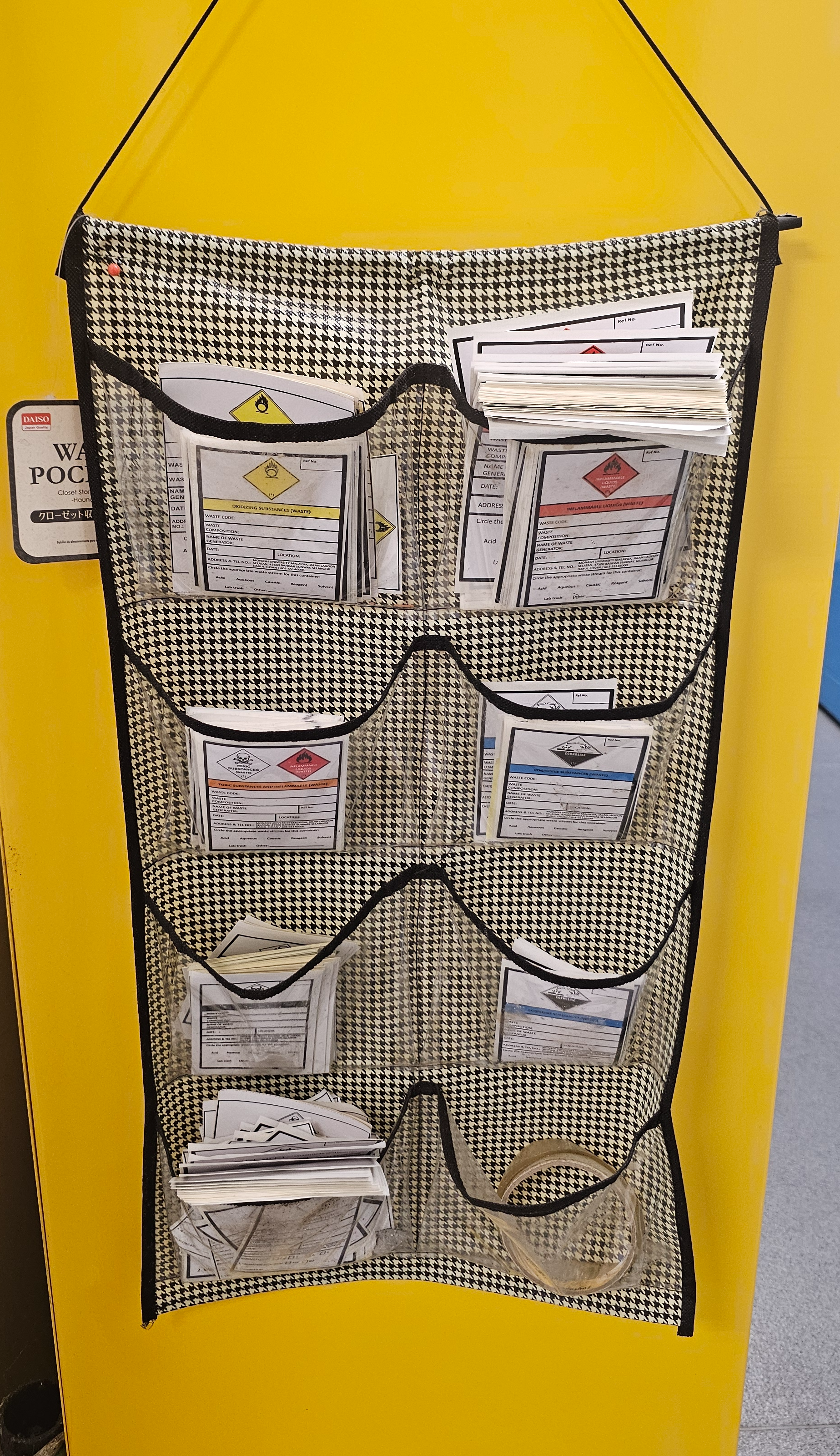 Pocket organiser hanging on the yellow flammables cabinet holding blank scheduled waste labels sorted by hazard class: oxidising, flammable liquids, toxic, corrosive