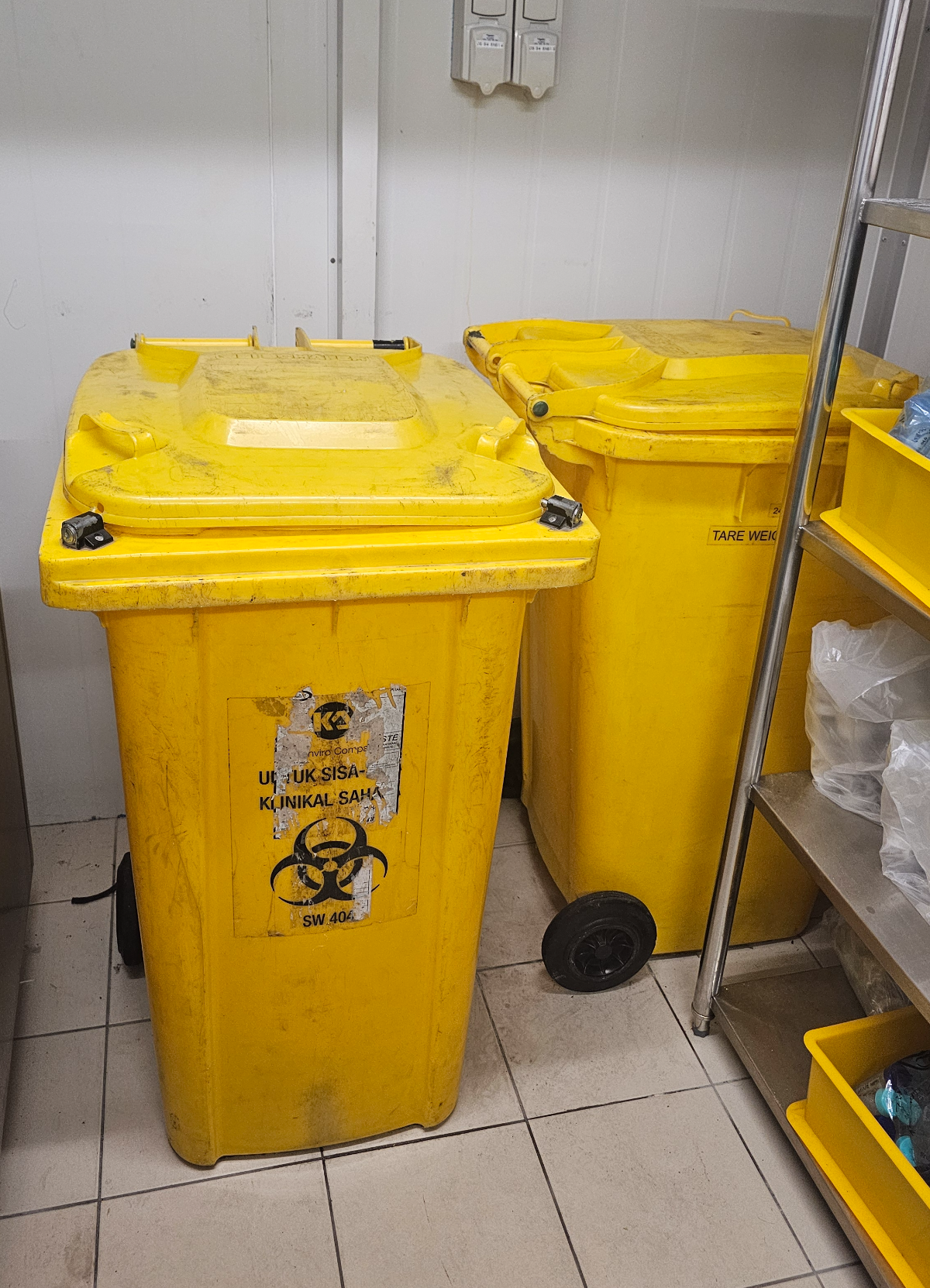 Two yellow SW 404 clinical waste bins inside the cold room, with a stainless steel rack beside them
